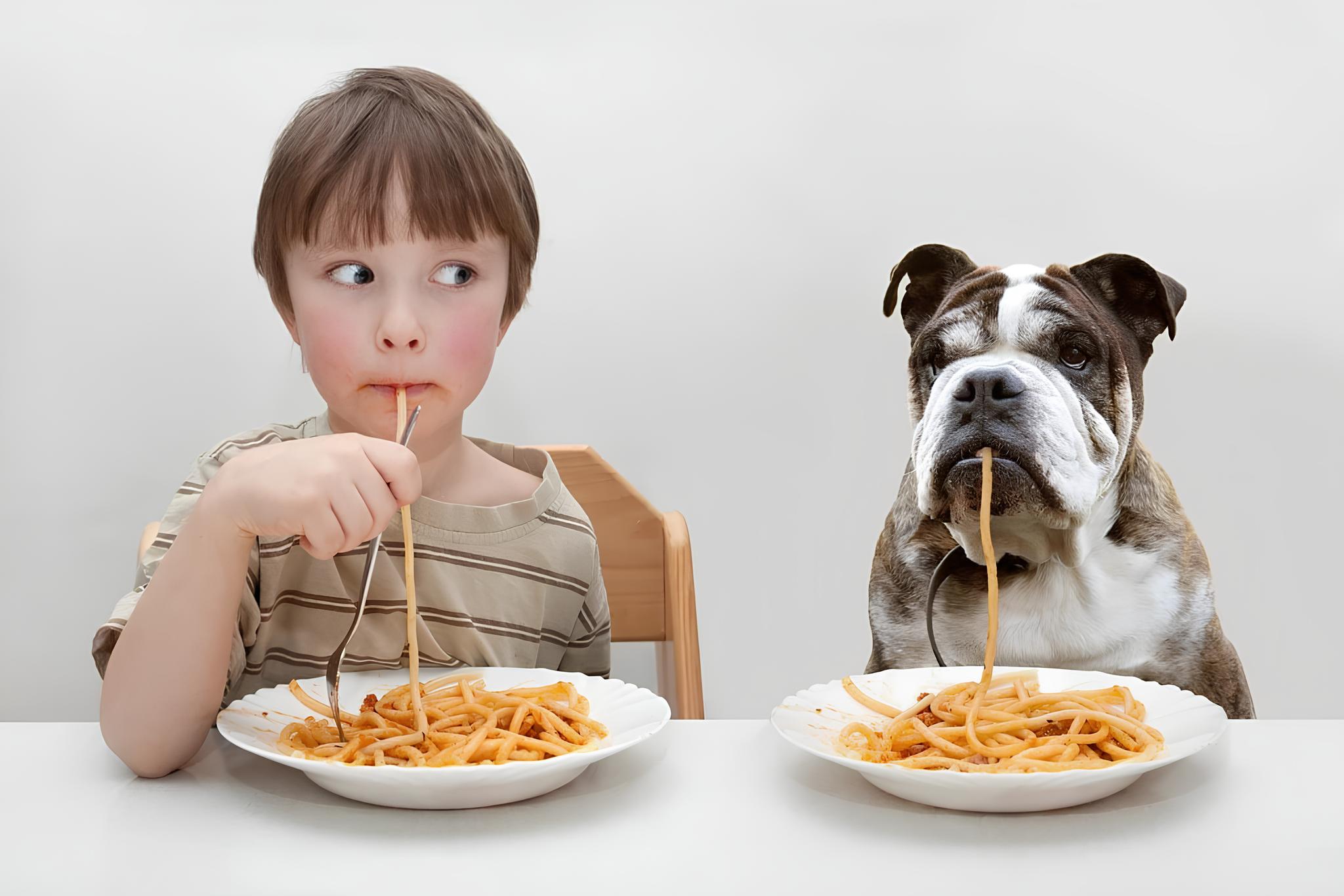 um criança comendo um prato de macarrão e ao seu lado um cachorro com seu prato também de macarrão.